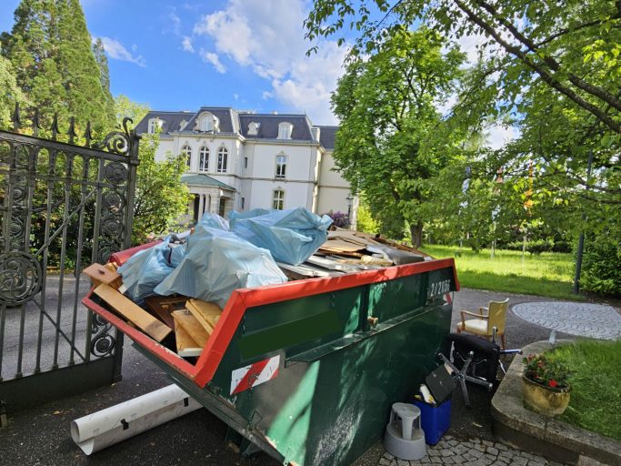 Ein Abfallcontainer mit Holz und Abdeckblau auf einem Grundstück mit einem großen Haus im Hintergrund